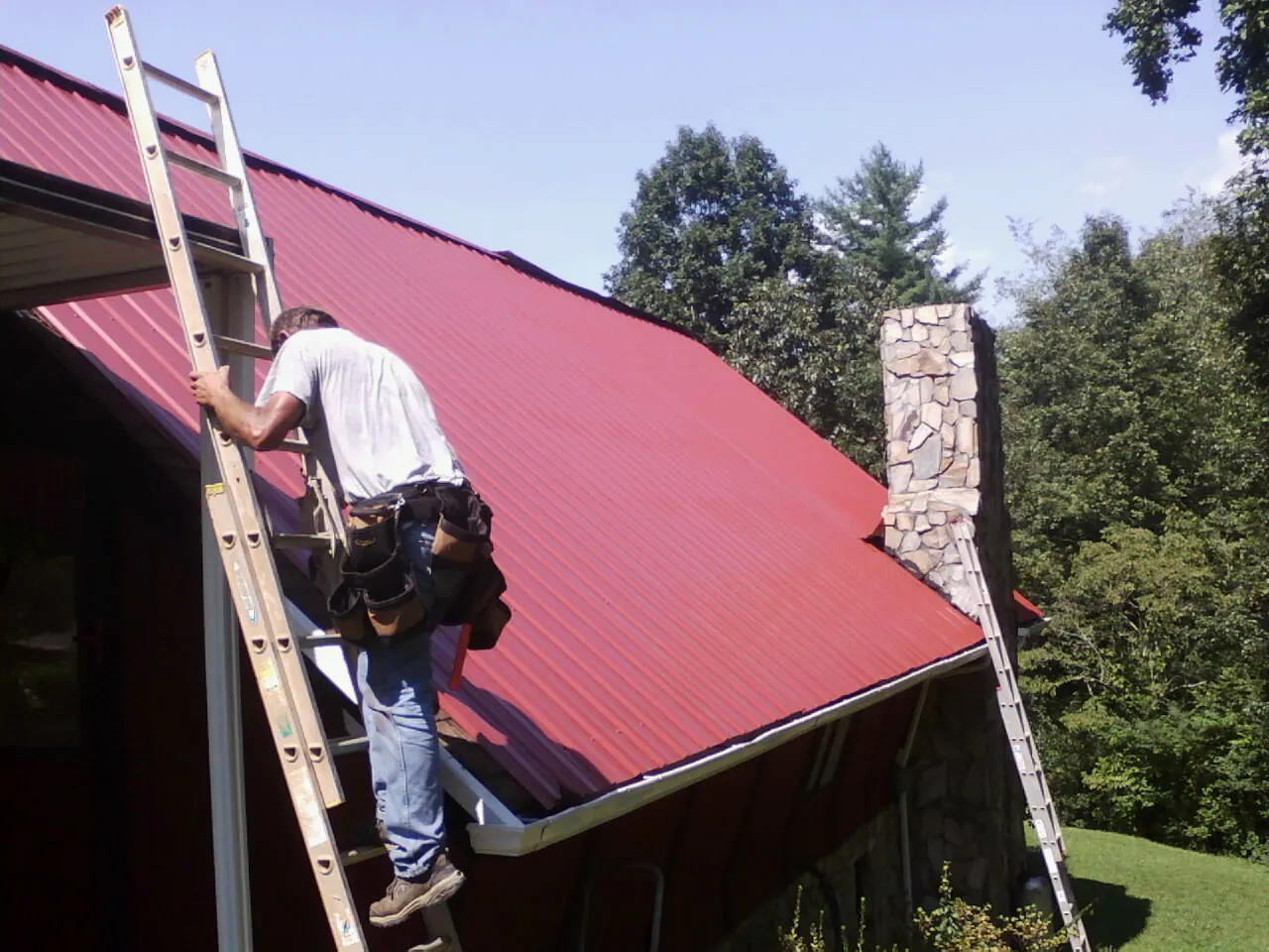 Roofer on a ladder beside a red metal roof and stone chimney — Soffit & Fascia Repair by Roofing Brothers in Havelock